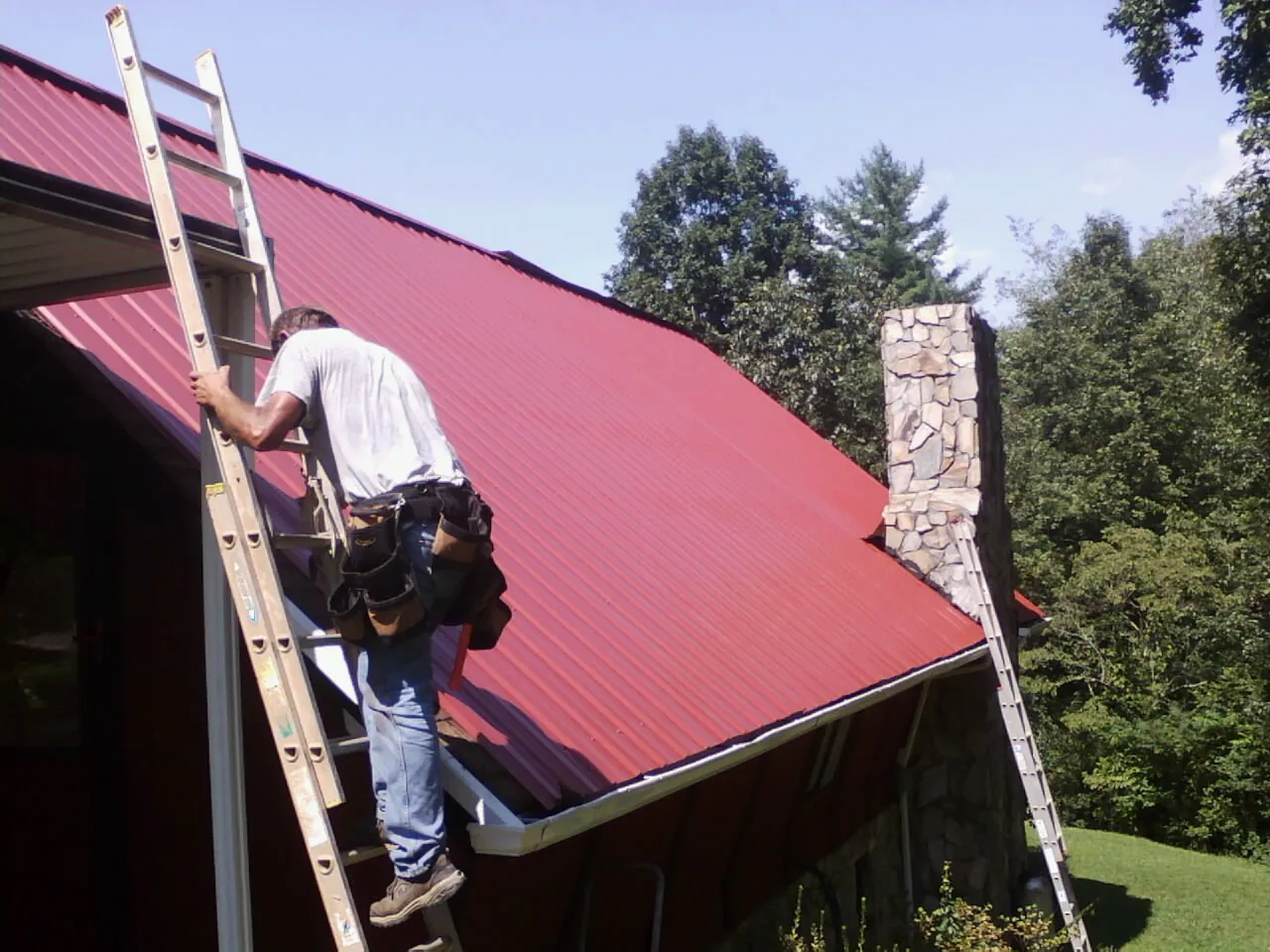 Roofer on a ladder beside a red metal roof and stone chimney — Soffit & Fascia Repair by Roofing Brothers in Newmarket